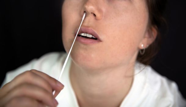 Young woman taking a Covid-19 SARS-CoV-2 test during Corona pandemic on dark black background, quick home test coronavirus closeup nose swab