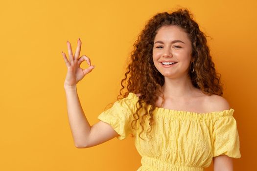 Young curly smiling woman showing ok sign against yellow background, close up