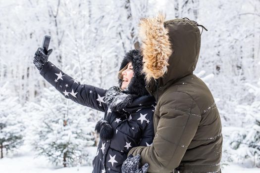 Technologies and relationship concept - Happy smiling couple taking a selfie in a winter forest outside.