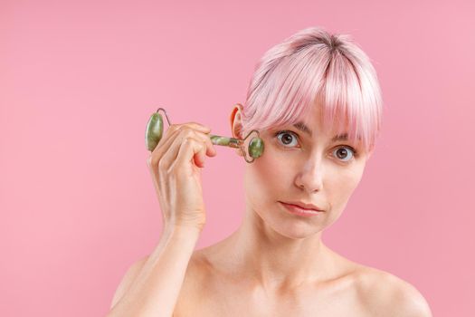 Surprised young woman with pink hair looking at camera while using natural jade facial roller for skin care isolated over pink background. Face massage and beauty trends concept