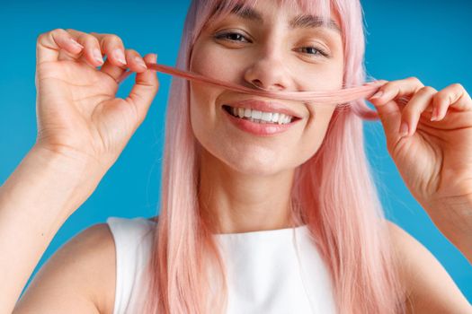 Close up portrait of happy young woman with natural long pink dyed hair holding a strand of hair as a moustache and smiling at camera, posing isolated over blue studio background. Hair care concept