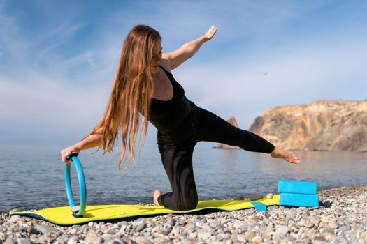 Young woman with long hair, fitness instructor in black Sportswear Leggings and Tops, stretching on a yoga mat with magic pilates ring near the sea on a sunny day, female fitness yoga routine concept.