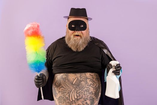 Happy emotional middle aged man with overweight in hero suit holds colorful dust brush and spray bottle standing on purple background in studio