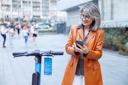 Pretty senior woman with glasses and mobile phone looks at e-scooter exposed on city street on autumn day. New transportation technology
