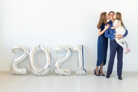 Young happy family mother and father and daughter standing near balloons shaped like numbers 2021 on white background. New year, Christmas and holiday.