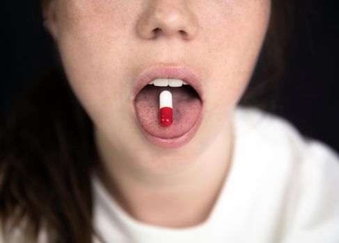 Young woman,teenager with Capsule on tong on dark black background, taking pills. medicine,drugs,health concept close up portrait