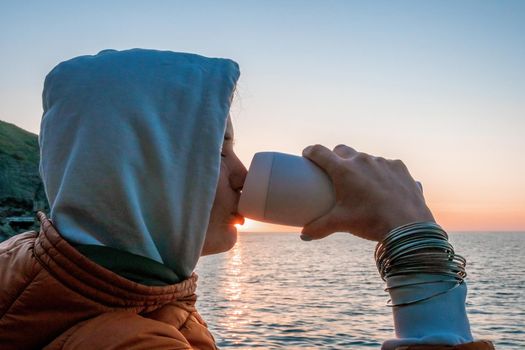 woman traveler drinks coffee with a view of the mountain landscape. A young tourist woman drinks a hot drink from a cup and enjoys the scenery in the mountains. Trekking concept