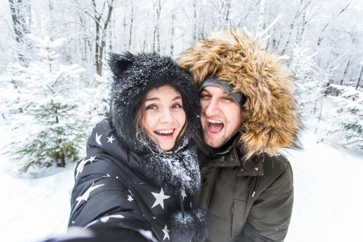 Technologies and relationship concept - Happy smiling couple taking a selfie in a winter forest outside.