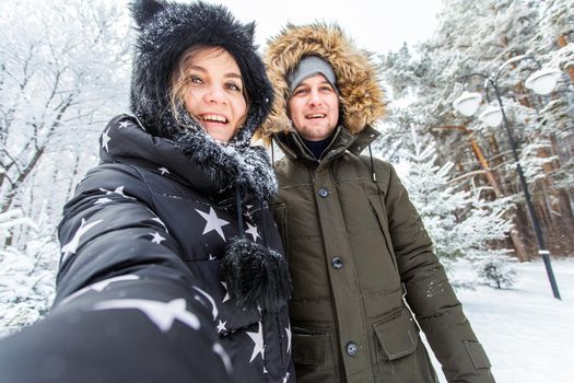 Technologies and relationship concept - Happy smiling couple taking a selfie in a winter forest outside.
