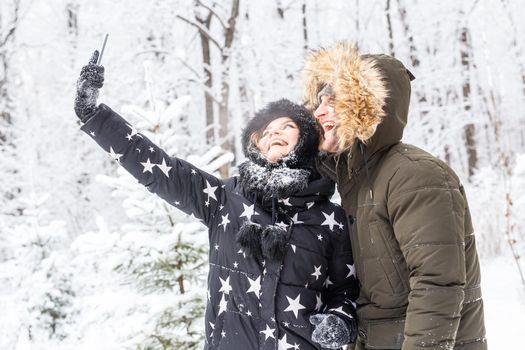 Technologies and relationship concept - Happy smiling couple taking a selfie in a winter forest outside.