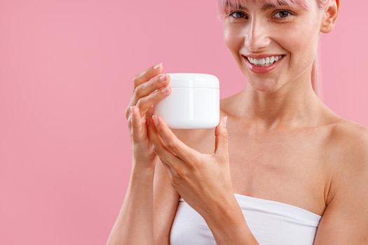 Cropped portrait of young woman smiling at camera, holding white jar with moisturizing body lotion after shower isolated over pink background. Beauty, spa, body care concept