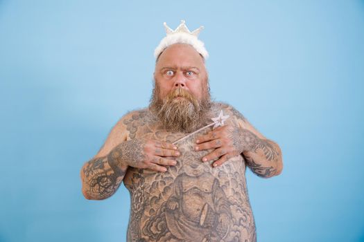 Funny surprised obese man with toy crown and magic stick covers breasts by hands shying on light blue background in studio