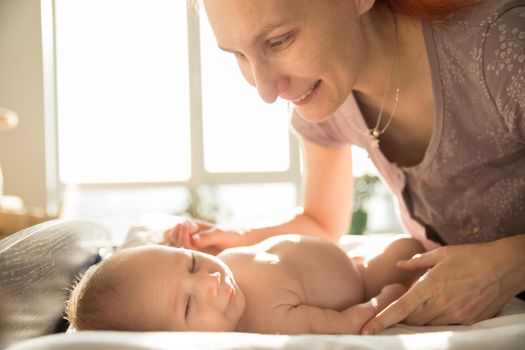 Smiling ginger mother takes care of her small baby lying on a bed. Mid shot
