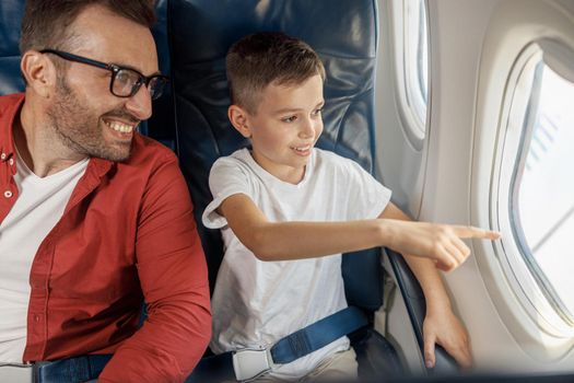 Happy little boy looking out the window, showing something to his father while sitting near the window, traveling by plane together. Family, transportation, vacation concept