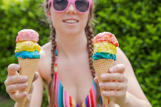 Beautiful girl holding two colorful ice cream with sprinkles wearing a rainbow colored swimsuit in the summer, pink sunglasses and braids happy vacation,holidays concept sunshine