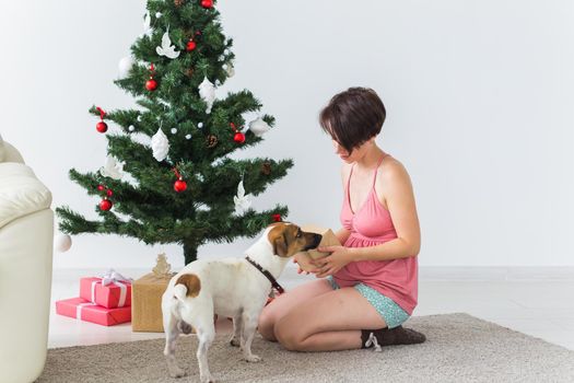 Happy woman with dog opening Christmas gifts. Christmas tree with presents under it. Decorated living room.