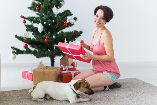 Happy woman with dog opening Christmas gifts. Christmas tree with presents under it. Decorated living room.