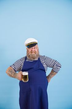 Confident bearded man with overweight in sailor suit with apron and captainhat holds mug of tasty beer on light blue background in studio