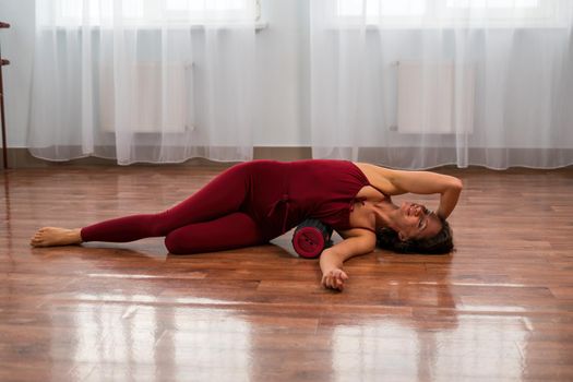 A young woman fitness instructor in red Sportswear Leggings and Top stretching in the gym before her pilates, on a yoga mat near the large window on a sunny day, female fitness yoga routine concept