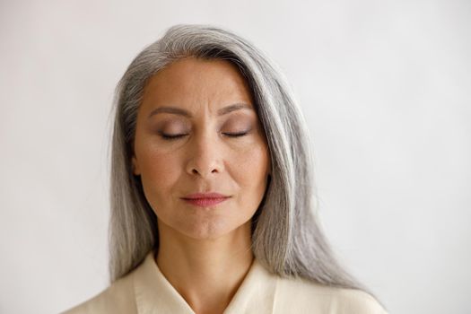 Pretty tranquil grey haired lady with closed eyes and elegant makeup poses on light background in studio. Mature beauty lifestyle