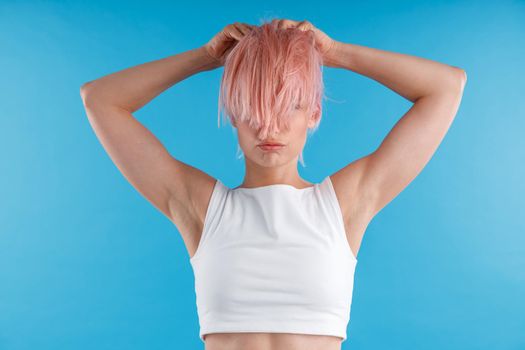 Young woman in white shirt playing with smooth straight pink hair ponytail, hiding her face behind it while posing isolated over blue studio background. Beauty, hair care concept