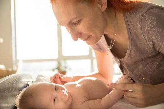 Smiling mother takes care of her small baby lying on a bed. Mid shot