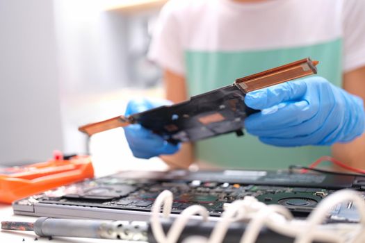 Gloved hands are holding a spare part from the computer, master plan. There is a broken laptop on the table. Electronic device repair service