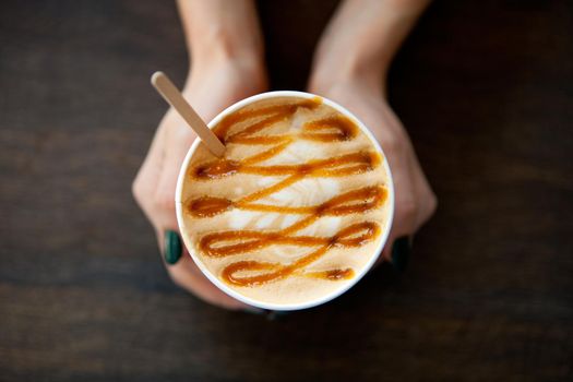 Caramel Big Coffee paper cup with milk in womans hand on the wooden table. cappuccino or latte drink, cup of coffee on table flat lay view. Cup of cafe au lait. Milk paintings. Coffee for a girl