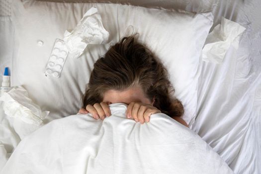 A sick young woman in bed, lying with her head under the blankets and a pile of tissues next to her top view. Fever staying home from work. white sheets and pillow