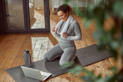 Jolly slim female is sitting on mat and looking at laptop while doing training at home