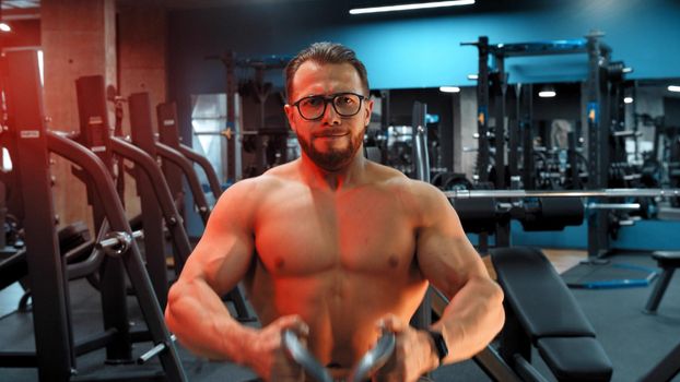 A bearded attractive man bodybuilder doing strength exercises in the sports gym - pulling the weight with his hands in neon lighting. Mid shot
