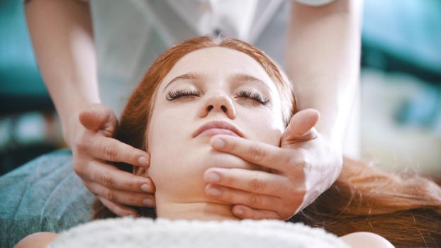 Massage - masseuse kneading the chin area to a young woman using her palms - indoor