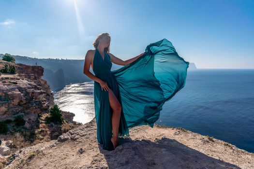 A girl with loose hair in a long mint dress descends the stairs between the yellow rocks overlooking the sea. A rock can be seen in the sea. Sunny path on the sea from the rising sun.