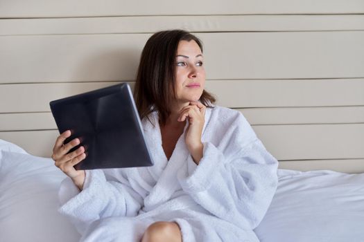 Dreaming mature woman in white bathrobe with digital tablet looking to the side, relaxing sitting in bed, copy space