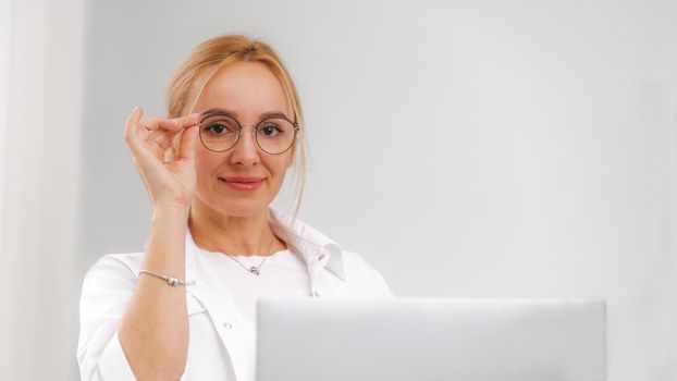 A blonde doctor in glasses or a cosmetologist in a white coat is sitting at a computer. She reads and types on the computer. Concept of medicine, beauty, cosmetologist, masseur