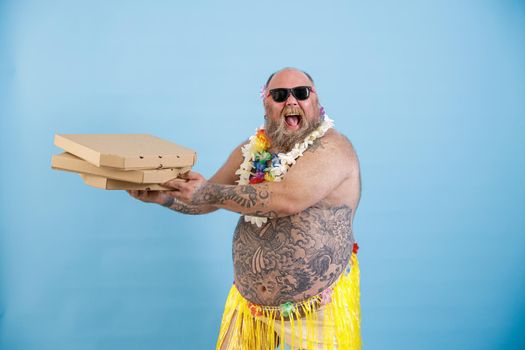 Happy obese man with sunglasses in yellow grass skirt and flowers garland holds boxes stack of pizza on light blue background in studio