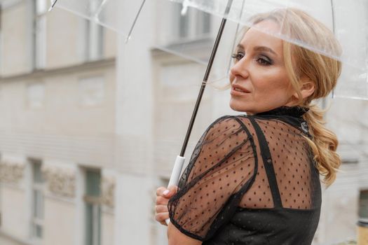 The blonde stands under a transparent umbrella during the rain. The fall season. Rear view. The woman is dressed in a black lace dress, her hair pulled back in a ponytail