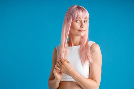 Young woman touching her smooth natural long pink dyed hair and looking surprised, posing isolated over blue studio background. Beauty, hair care concept