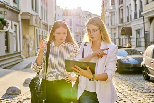 Two young businesswomen walking along city street, talking and looking in screen of digital tablet. Business, people, technology, teamwork, communication, lifestyle concept