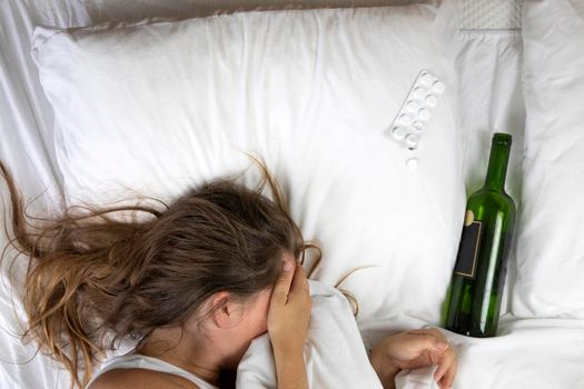 Sleeping drunk girl with empty wine bottle. Lying on the bed fainted, alcohol abuse,addiction concept top view white sheets