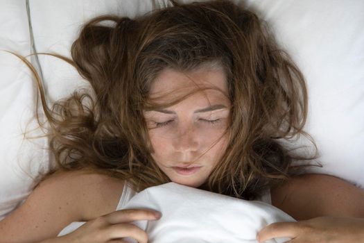 Beautiful young woman sleeping in bed with messy brown hair top view, lying with white bed sheets and freckles close up