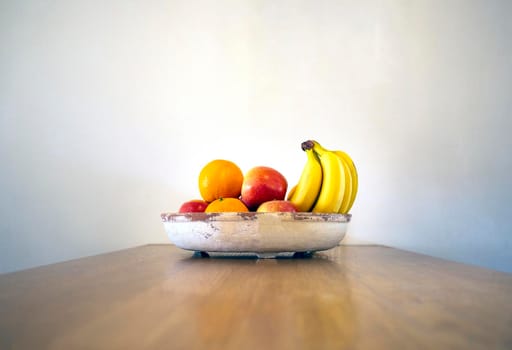Healthy fresh fruit bowl on wooden table with Bananas, oranges and apples near white wall modern design on wooden table, copy space healthy still life front view home decoration