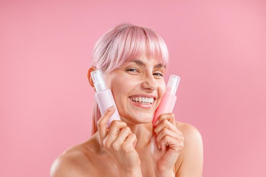 Pleased young woman with pink hair holding two plastic spay bottles with beauty products near her face, posing isolated over pink background. Beauty, spa, body care concept