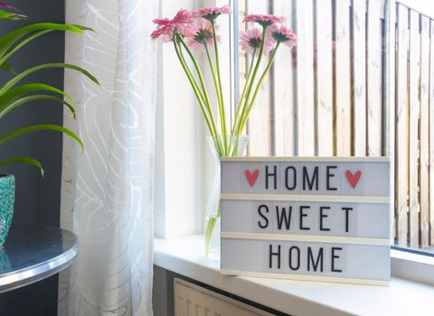 Sign Home sweet home text on lightbox ,windowsill near window with pink flowers, decorative frame modern interior background, stylish house cozy corner in lovely living room summer