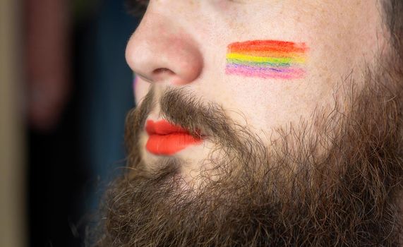 Young handsome bearded man with pride flag on his cheek, rainbow flag standing for LGBTQ, Gender right and sexual minority. Portrait closeup