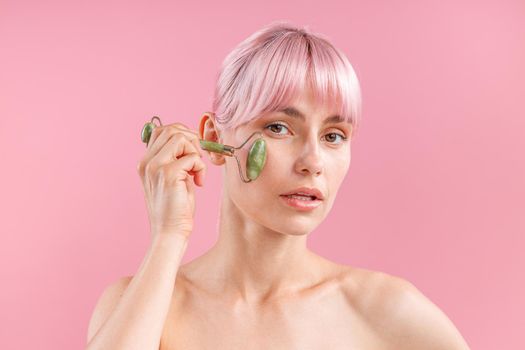 Portrait of young naked woman with pink hair using natural jade facial roller for skin care, posing isolated over pink background. Face massage and beauty trends concept