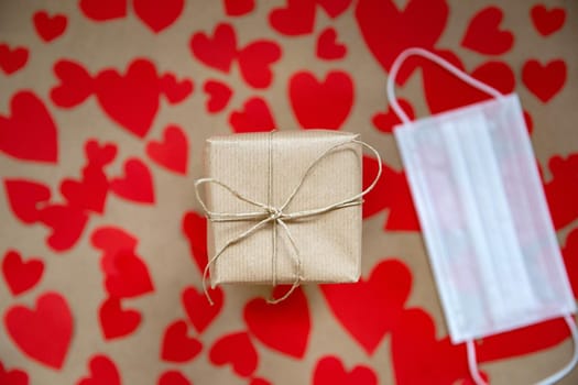 Female hands holding gift box for Valentines Day wrapped in Craft paper top view, romantic red hearts and Medical mask on background for Coronavirus, Covid-19 and Holliday, Valentine, Birthday concept. copy space