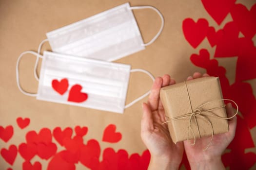 Female hands holding gift box for Valentines Day wrapped in Craft paper top view, romantic red hearts and Medical mask on background for Coronavirus, Covid-19 and Holliday, Valentine, Birthday concept. copy space