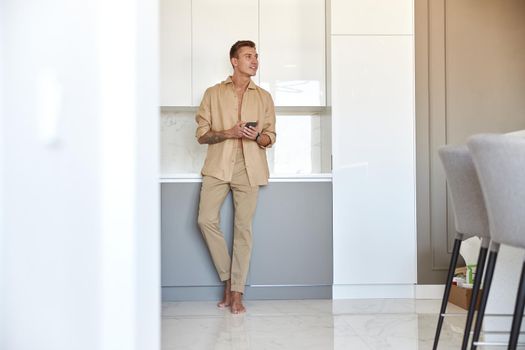 Hadsome smiling man is chilling on the minimalistic kitchen with smartphone