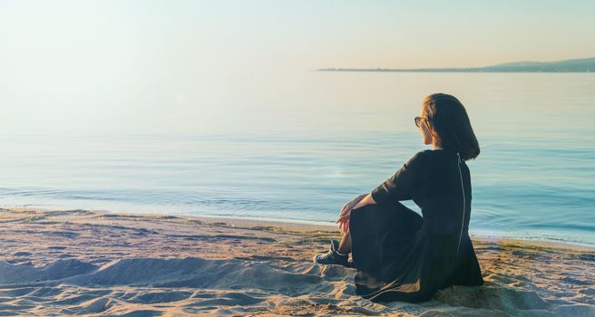 Beautiful young woman in black dress and sneakers sitting on beach and enjoying view of sea at sunny day